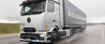 Der Mercedes-Benz NextGenH2 fährt auf einer Teststrecke durch eine Steilkurve. Die silbern lackierte Sattelzugmaschine zieht einen KOfferauflieger