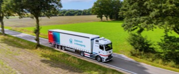 Der eActros 600 von Mercedes Benz im Branding des Logistkdienstleisters Fiege fährt auf einer Straße durch eine Parklandschaft mit grünen und braunen Äckern sowie Bäumen und im Hintergrund ein Wald. Auf dem Trailer steht neben dem Fiege-Logo auf einem Farbhintergrund im rot-blau und türkisen Farbspektrum auf weißem Hintergrund neben dem Symbol einer Ladesäule "Elektrisch in Bewegeung."