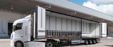 Seitliche Frontansicht der B.BO Obensider von Schmitz Cargobull. Die Seitenwände auf der linken Seite sind geöffnet. Das Fahrzeug parkt auf einem Betriebshof, im Hintergrund sind viele Laderaupen sichtbar