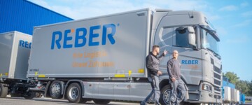 Zwei Männer stehen vor einem Lkw der Reber Logistik,der vor einer Lagerhalle parkt