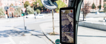 Im Bild der Blick nach rechts aus dem Innenraum des Fahrerhauses im Lkw mit Fokus auf einen Abbiegeassistenten von Mercedes-Benz, der grade einen E-Scooter Fahrer neben dem Lkw anzeigt und über ein kleines rotes Warndreieck auf die Gefahr aufmerksam macht. Der Abbiege-Assistent von Mercedes-Benz ist laut Hersteller in der Lage, ein sich bewegendes oder stehendes Objekt in der Überwachungszone auf der rechten Seite des Lkw zu erkennen