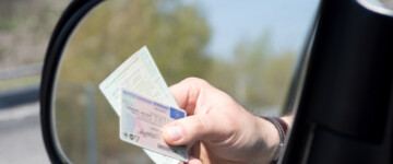 Im Bild sieht man in einem Rückspiegel eine Hand, die Führerschein und Fahrzeugpapiere aus dem Fenster hält, möglicherweise im Rahmen einer Kontrolle