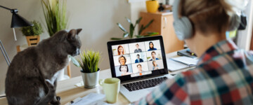 Eine junge Frau sitzt vor ihrem Laptop in einer Videokonferenz mit Kollegen im Homeoffice. Neben ihr auf dem Schreibtisch sitzt eine Katze (Symbolbild mit Fotomodell)