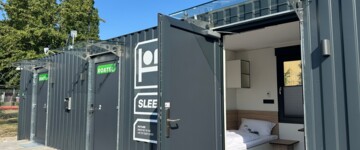 Am Lagerstandort Gimbsheim von TST in Rheinland-Pfalz ist ein Mikrohotel für Berufskraftfahrer des Anbieters Roatel entstanden. Sie können dort ihre regelmäßige wöchentliche Ruhezeit verbringen und haben zugleich einen Stellplatz für den Lkw in der Nähe. Zu sehen ist der schwarze Überseecontainer, der die vier kleinen Apartments beherbergt. Eine offene Tür ermöglicht den Blick auf das Bett, ein Nachttischchen und Regalfächer.