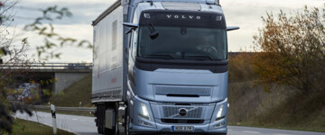 Ein Volvo FH Aero I-Save fährt bei bedecktem Himmel und schummerigen Lichtverhältnissen auf einer Straße durch die Landschaft, von vorne aufgenommen, zu sehen ist eine buschbestandene Böschung und im Hintergrund eine Brücke