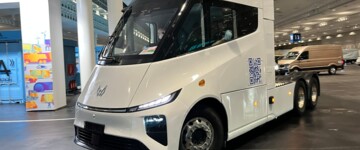 Der E-Lkw fährt in einer Lagerhalle. Die weiß lackierte Dreiachs-Sattelzugmaschine ist ohne Auflieger unterwegs 