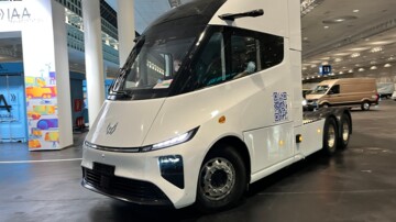 Der E-Lkw fährt in einer Lagerhalle. Die weiß lackierte Dreiachs-Sattelzugmaschine ist ohne Auflieger unterwegs 