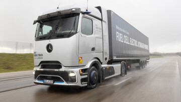 Der Mercedes-Benz NextGenH2 fährt auf einer Teststrecke durch eine Steilkurve. Die silbern lackierte Sattelzugmaschine zieht einen KOfferauflieger