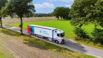 Der eActros 600 von Mercedes Benz im Branding des Logistkdienstleisters Fiege fährt auf einer Straße durch eine Parklandschaft mit grünen und braunen Äckern sowie Bäumen und im Hintergrund ein Wald. Auf dem Trailer steht neben dem Fiege-Logo auf einem Farbhintergrund im rot-blau und türkisen Farbspektrum auf weißem Hintergrund neben dem Symbol einer Ladesäule "Elektrisch in Bewegeung."