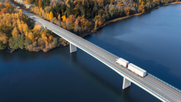 Lkw fährt auf Brücke über einen Fluss in Richtung eines herbstlichen Waldes