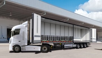 Seitliche Frontansicht der B.BO Obensider von Schmitz Cargobull. Die Seitenwände auf der linken Seite sind geöffnet. Das Fahrzeug parkt auf einem Betriebshof, im Hintergrund sind viele Laderaupen sichtbar