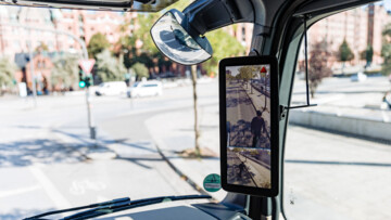 Im Bild der Blick nach rechts aus dem Innenraum des Fahrerhauses im Lkw mit Fokus auf einen Abbiegeassistenten von Mercedes-Benz, der grade einen E-Scooter Fahrer neben dem Lkw anzeigt und über ein kleines rotes Warndreieck auf die Gefahr aufmerksam macht. Der Abbiege-Assistent von Mercedes-Benz ist laut Hersteller in der Lage, ein sich bewegendes oder stehendes Objekt in der Überwachungszone auf der rechten Seite des Lkw zu erkennen