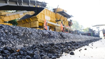 Asphaltreste liegen neben der Autobahn 215 zwischen dem Autobahndreieck Bordesholm (Schleswig-Holstein) und der Anschlussstelle Blumenthal. Im Hintergrund arbeiten ein Asphaltaufbringer und Arbeiter. Auf dem Teilstück wurde 2021 Kompaktasphalt aufgetragen. Dabei werden Binder- und Deckschicht in einem Arbeitsgang und auf voller Fahrbahnbreite eingebaut, was Einfluss auf Qualität und Lebensdauer des Belags hat.