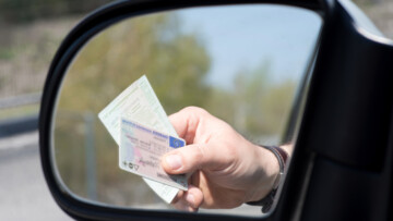 Im Bild sieht man in einem Rückspiegel eine Hand, die Führerschein und Fahrzeugpapiere aus dem Fenster hält, möglicherweise im Rahmen einer Kontrolle