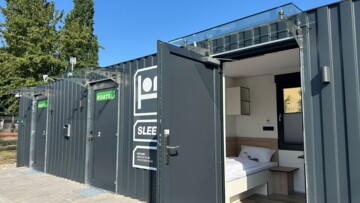 Am Lagerstandort Gimbsheim von TST in Rheinland-Pfalz ist ein Mikrohotel für Berufskraftfahrer des Anbieters Roatel entstanden. Sie können dort ihre regelmäßige wöchentliche Ruhezeit verbringen und haben zugleich einen Stellplatz für den Lkw in der Nähe. Zu sehen ist der schwarze Überseecontainer, der die vier kleinen Apartments beherbergt. Eine offene Tür ermöglicht den Blick auf das Bett, ein Nachttischchen und Regalfächer.