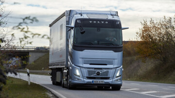 Ein Volvo FH Aero I-Save fährt bei bedecktem Himmel und schummerigen Lichtverhältnissen auf einer Straße durch die Landschaft, von vorne aufgenommen, zu sehen ist eine buschbestandene Böschung und im Hintergrund eine Brücke