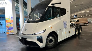 Der E-Lkw fährt in einer Lagerhalle. Die weiß lackierte Dreiachs-Sattelzugmaschine ist ohne Auflieger unterwegs 