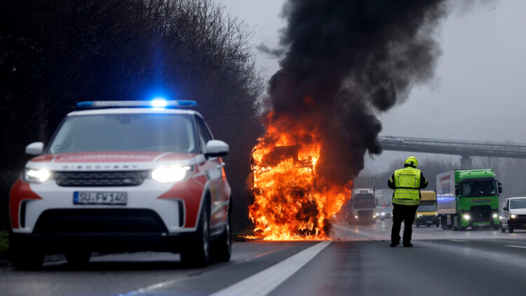 Ein brennender Lkw steht auf einer Autobahn, im Vordergrund Sicherheitskräfte