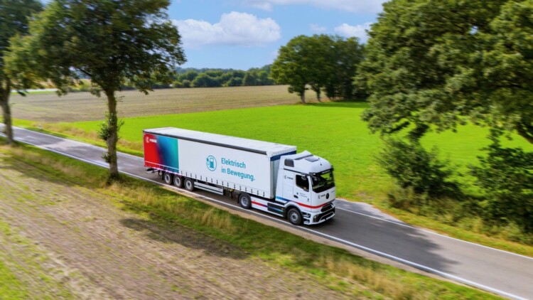 Der eActros 600 von Mercedes Benz im Branding des Logistkdienstleisters Fiege fährt auf einer Straße durch eine Parklandschaft mit grünen und braunen Äckern sowie Bäumen und im Hintergrund ein Wald. Auf dem Trailer steht neben dem Fiege-Logo auf einem Farbhintergrund im rot-blau und türkisen Farbspektrum auf weißem Hintergrund neben dem Symbol einer Ladesäule "Elektrisch in Bewegeung."
