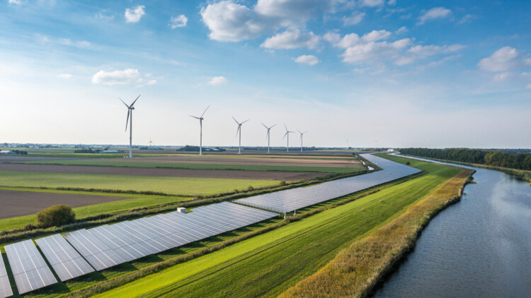 neben einem Fluss zieht sich eine Reihe Solaranlagen durchs Land, im Hintergrund Windräder