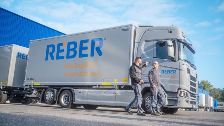 Zwei Männer stehen vor einem Lkw der Reber Logistik,der vor einer Lagerhalle parkt