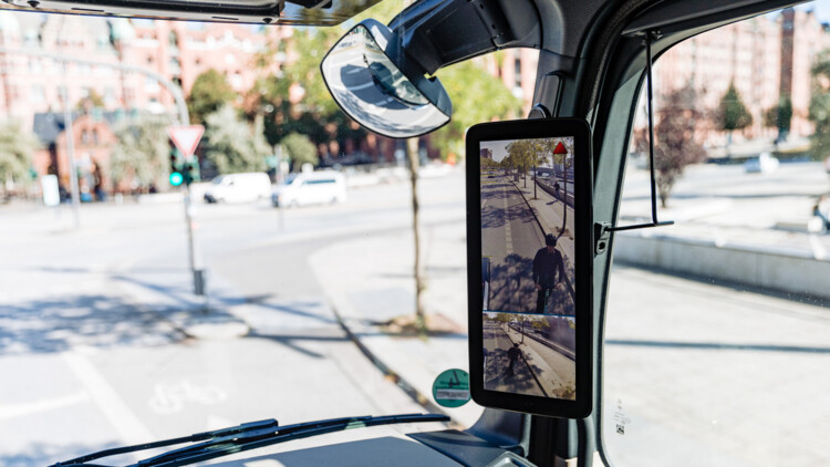 Im Bild der Blick nach rechts aus dem Innenraum des Fahrerhauses im Lkw mit Fokus auf einen Abbiegeassistenten von Mercedes-Benz, der grade einen E-Scooter Fahrer neben dem Lkw anzeigt und über ein kleines rotes Warndreieck auf die Gefahr aufmerksam macht. Der Abbiege-Assistent von Mercedes-Benz ist laut Hersteller in der Lage, ein sich bewegendes oder stehendes Objekt in der Überwachungszone auf der rechten Seite des Lkw zu erkennen