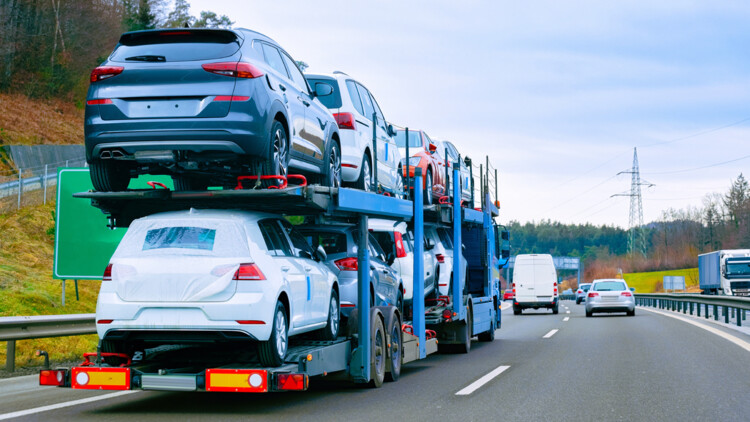 Ein Lkw-Autotransporter fährt auf einer Autobahn, Aufnahme von seitlich schräg hinten mit Blick auf weitere Fahrzeuge, die vor dem Lkw fahren sowie einen normalen Lkw auf der Gegenfahrbahn