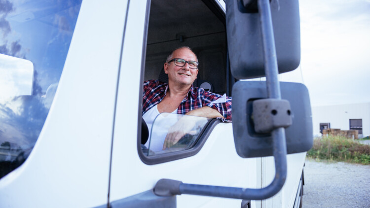 Älterer Mann oder Renter sitzt hinter dem Steuer eines Lkw und schaut lächelnd aus dem offenen Fenster in den Seitenspiegel. 