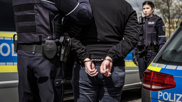 Zwei Polizisten verhaften einen Kriminellen. Im Bildvordergrund stehen zwischen zwei Polizeifahrzeugen ein Polizist und der Verbrecher mit Handschellen mit dem Rücken zum Betrachter. Im Hintergrund steht eine Polizistin und blickt ihrem Kollegen und dem Verhafteten entgegen.