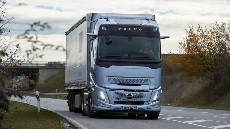 Ein Volvo FH Aero I-Save fährt bei bedecktem Himmel und schummerigen Lichtverhältnissen auf einer Straße durch die Landschaft, von vorne aufgenommen, zu sehen ist eine buschbestandene Böschung und im Hintergrund eine Brücke